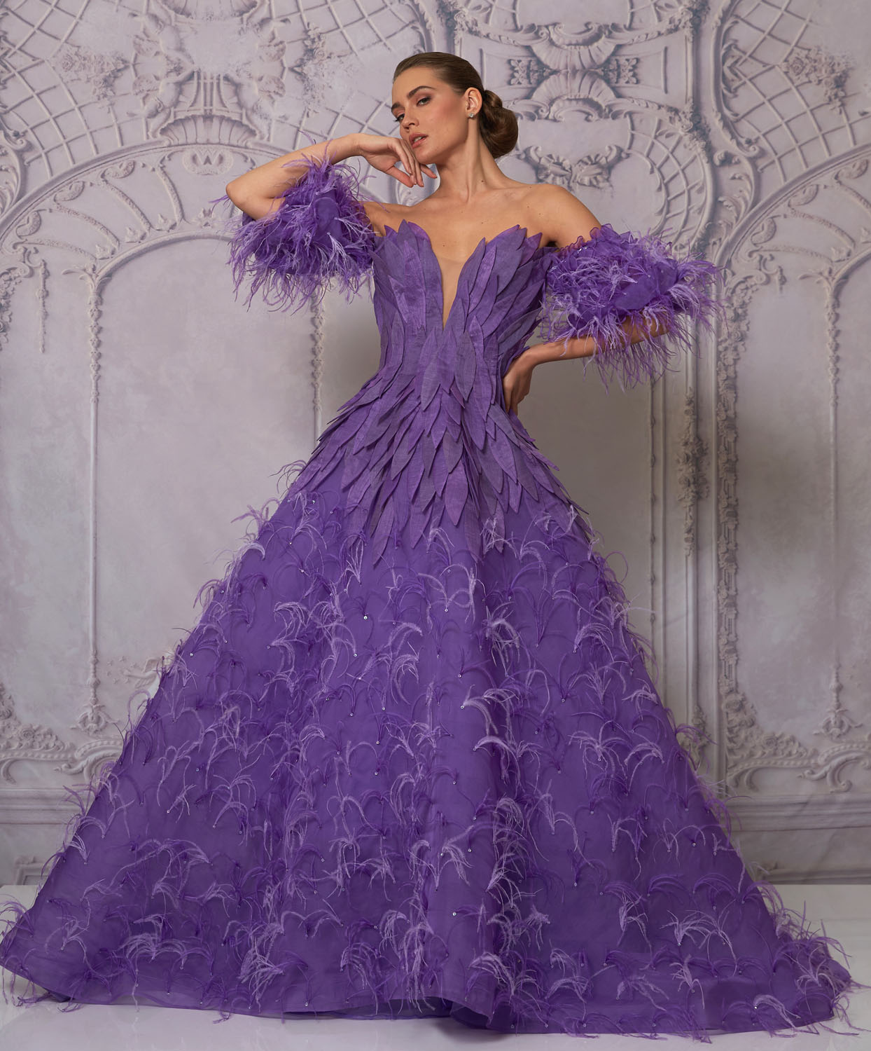 A full-length image of a woman wearing an off-the-shoulder, vibrant purple ball gown. The dress features a deep V neckline with layered petal-like fabric details on the bodice. The voluminous skirt is adorned with delicate feather-like embellishments throughout., image 1